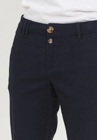 Marineblå bukser med en glat tekstur, der har to messingknapper, zip-fly og standard bæltesløjfer for justerbar pasform.