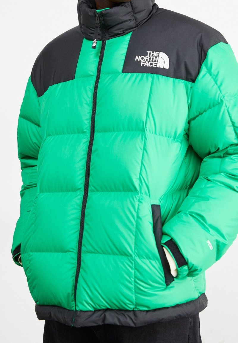 Světlý zelený péřový kabát s černými akcenty, vybavený vysokým límcem, předním zipem a polstrovanými oblastmi pro izolaci. Logo na hrudi.