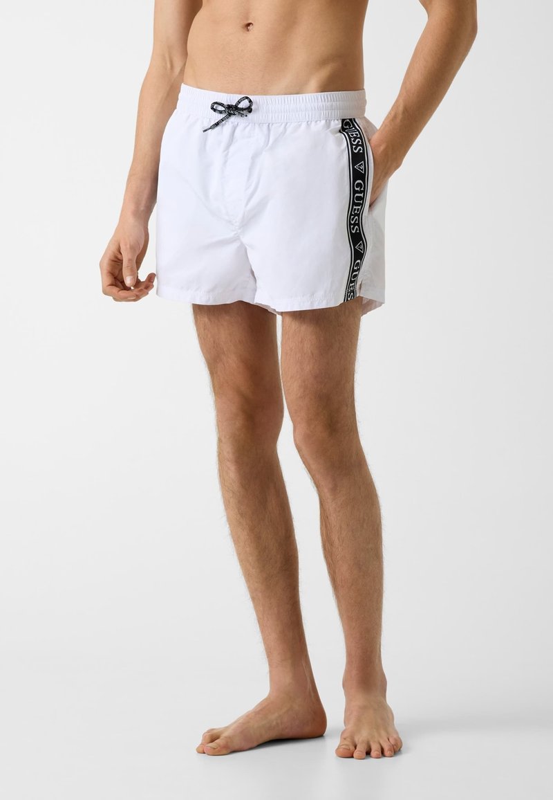 Homme portant un short de bain blanc avec une bande noire du logo Guess sur les côtés, debout pieds nus devant un fond uni.