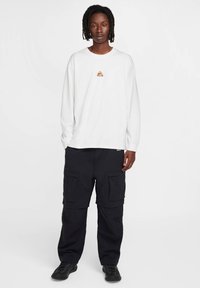 Cămașă albă cu mâneci lungi, având un mic logo portocaliu, asortată cu pantaloni cargo negri, cu multiple buzunare și o croială relaxată. Încălțăminte neagră.