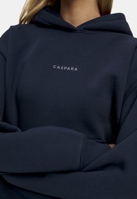 Marineblå hettegenser laget av mykt stoff med kengurulomme, ribbestrikkede ermer og en liten hvit "CASPARA"-logo på brystet.