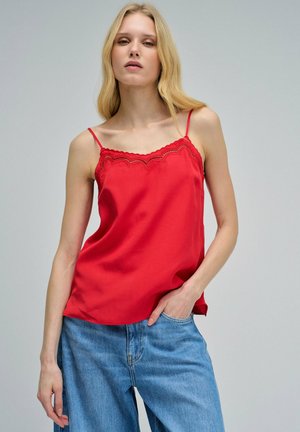 Femme blonde portant un haut sans manches rouge orné de dentelle et un jean en denim bleu clair, une main dans la poche, sur fond uni.