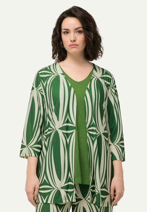 Giacca kimono verde a fantasia con maniche 3/4 sopra una maglia verde a tinta unita. Il tessuto presenta un audace motivo a foglie nei colori verde e crema.