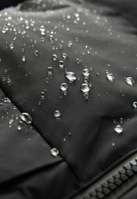 Tissu noir résistant à l'eau avec des gouttelettes visibles à la surface, présentant une finition texturée et une fermeture éclair sur le bord.