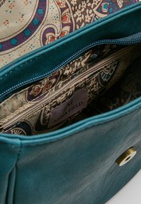 Wnętrze torby z niebieskozielonej sztucznej skóry z wzorzystą tkaniną podszewką, wyposażone w kieszeń na zamek i metkę z napisem "ANNA FIELD".