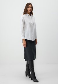 Witte langslijf knoopshirt met lange mouwen, zachte stof, gebogen zoom, gecombineerd met een donkergrijze knielange rok en zwarte hoge laarzen met hak.