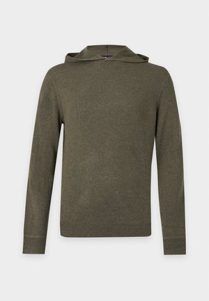 Jeune homme portant un sweat à capuche vert olive, un jean bleu ample et des baskets blanches, debout devant un fond blanc uni.