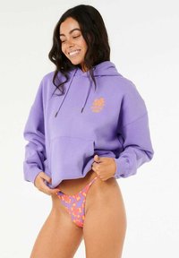 Paarse cropped hoodie met een trekkoordkap en een voorzak, gedragen met een roze en oranje gebloemd bikinionderdeel. Zacht materiaal met een relaxed fit.