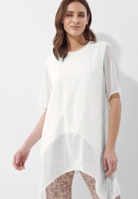 Witte tuniek met korte mouwen en een vloeiend, doorschijnend onderkleed. Heeft een ronde halslijn en wordt gedragen over lichtgekleurde, gemusterde leggings.