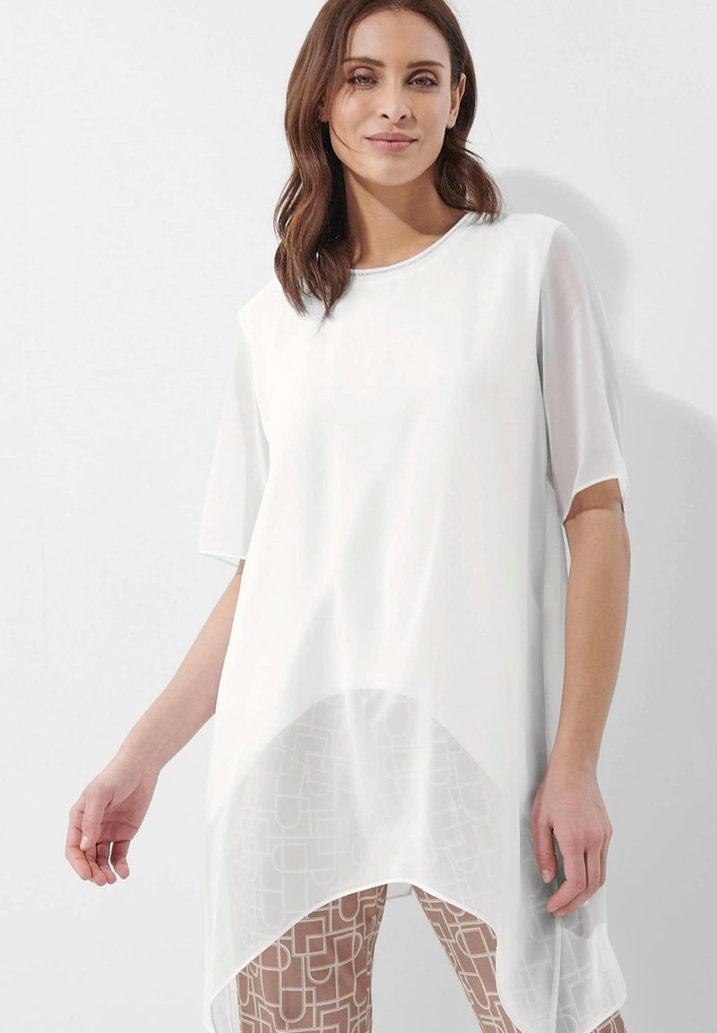 Witte tuniek met korte mouwen en een vloeiend, doorschijnend onderkleed. Heeft een ronde halslijn en wordt gedragen over lichtgekleurde, gemusterde leggings.