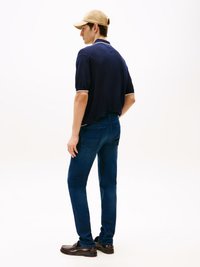 Camisola polo azul-marinho com detalhes brancos, calças de ganga escuras de corte justo e mocassins castanhos. O modelo também usa um boné de basebol bege.