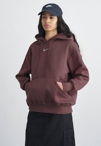 Νεαρό άτομο με μπορντό φούτερ Nike και μαύρο καπέλο Nike, με τα χέρια στην τσέπη του φούτερ, στέκεται μπροστά σε απλό ανοιχτό φόντο.