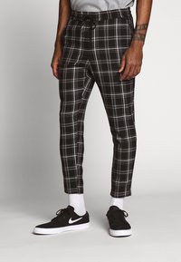 Man in zwart-wit geruite broek, witte sokken en zwarte Nike sneakers, staande tegen een effen lichte achtergrond.