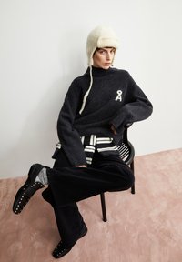 Dark gray chunky knit sweater with high neck and white logo, layered with striped shirt, black wide-leg corduroy pants, and black shoes with studs.