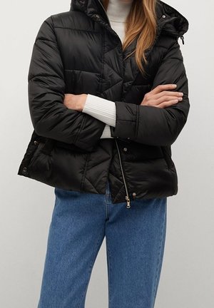 Femme portant une doudoune noire matelassée avec capuche, un pull en maille blanc côtelé et un jean bleu, les bras croisés sur la poitrine.