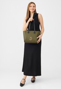 Olijfgroene totebag met zwarte handvatten, voorzien van een gouden logo en een subtiel ontwerp. Model draagt een zwarte geribbelde jurk en platte schoenen.