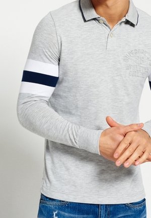 Mann trägt ein hellgraues langärmeliges Polo mit weißen und marineblauen Streifen am linken Arm, hält die Hände vor sich verschränkt, blaue Jeans sichtbar.