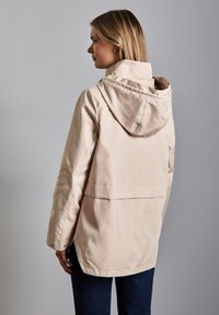 Veste imperméable beige avec capuche, empiècement au dos et détails à pression. Manches longues et coupe décontractée. Texture de tissu lisse.