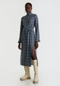Blumenkleid in Dunkelblau mit langen Ärmeln und hohem Kragen. Hat eine taillierte Taille und einen seitlichen Schlitz; kombiniert mit beigen kniehohen Stiefeln.