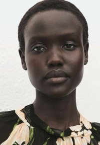 Primi piano di una persona con capelli corti e scuri e una pelle liscia, che indossa una blusa nera a fiori con dettagli gialli e bianchi su uno sfondo chiaro.