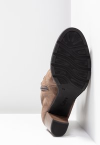 Botte en daim marron avec fermeture éclair latérale, semelle texturée noire, talon bloc, présentée semelle visible contre un fond blanc en angle.