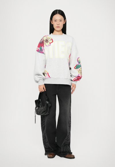 Gray sweatshirt with floral patterns and "STIEG" text, paired with black wide-leg jeans featuring side detailing. Model holds a black beaded bag.