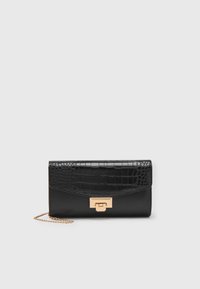 Pochette noire avec rabat à texture croco, fermoir rectangulaire doré et fine chaîne dorée sur fond blanc.