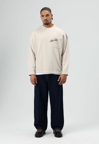 Licht beige oversized sweatshirt met een voorzak, voorzien van zwart geborduurde tekst. Gecombineerd met donkerblauwe wijde jeans en zwarte schoenen.