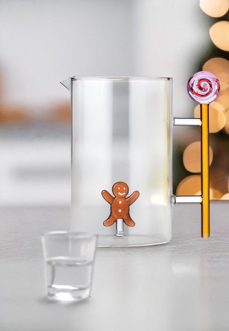 Przezroczysty dzbanek z motywem piernika wewnątrz, żółta rączka i różowy spiralny akcent. Obok znajduje się mała szklanka.