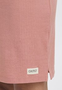 Roza teksturirana tkanina suknje s suptilnim mrežastim uzorkom. Ima prorez sa strane i ušivenu oznaku s natpisom "OXMO" u crnim slovima.