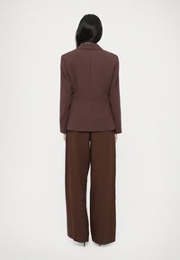 Hnedé na mieru ušité sako s padnutým pásom a široké hnedé nohavice, vyrobené z hladkej, štruktúrovanej látky. Pohľad zozadu.