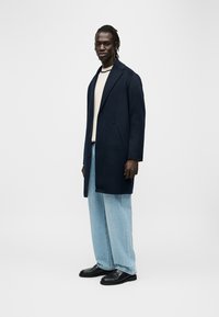 Manteau en laine bleu marine avec revers à encoche, deux poches avant, porté sur un pull beige et un jean large bleu clair, assorti à des mocassins noirs.