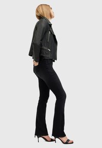 Svart läderbikerjacka med silverdragkedjor, svarta figurnära jeans och svarta öppna klackar, sedd från sidan mot en ljus bakgrund.