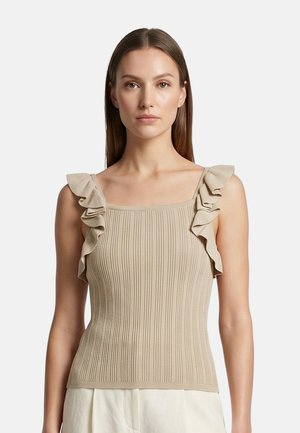 Donna con capelli castani lisci indossa un top beige a coste senza maniche con spalline arricciate e pantaloni bianchi su sfondo semplice.