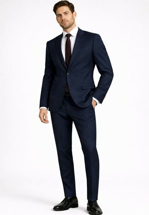 Homme debout portant un costume bleu marine foncé ajusté avec une chemise blanche, une cravate bordeaux et des chaussures habillées noires sur un fond uni.