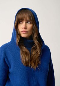 Blå huva med en strukturerad stickad mönster och hög krage. Har en avslappnad passform och långa ärmar, vilket betonar en avslappnad stil.