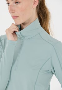 Pitkähiha, vaaleansininen urheilupaita, jossa on puolivetoketju kaula-aukossa, sileä rakenne ja vartalonmyötäinen muotoilu; sisältää hienovaraisia ompeluyksityiskohtia.