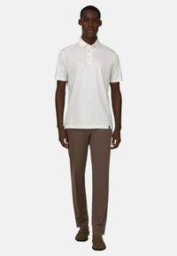 Homme debout, face à l'avant, portant un polo blanc à manches courtes, un pantalon marron et des mocassins en suède marron sur un fond uni.