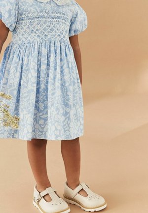 Robe à imprimé floral bleu clair avec manches bouffantes et corsage smocké, associée à des chaussures crème à bride en T avec détails perforés.