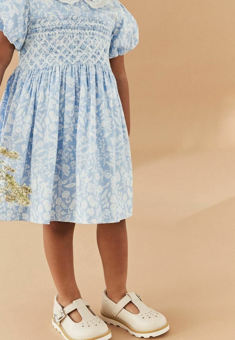 Robe à imprimé floral bleu clair avec manches bouffantes et corsage smocké, associée à des chaussures crème à bride en T avec détails perforés.