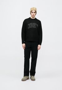 Pulover negru tricotat cu literele „OBEY”, asortat cu pantaloni negri și ghete verzi până la gleznă. Căciulă bej cu pete maro.