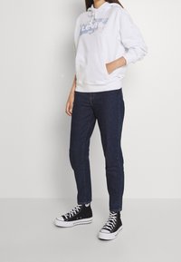 Bílá mikina s kapucí a modrým logem Levi's, přední klokaní kapsa, v kombinaci s tmavými denimovými džíny a černými vysokými teniskami s bílou podrážkou.