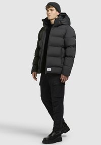 Sort puffjakke med hætte, som har lynlås og markante quiltede sektioner. Bæres over en sort sweater og kombineres med cargo-bukser.