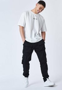Camiseta blanca de manga corta con logo, combinada con pantalones cargo negros que tienen múltiples bolsillos y zapatillas blancas. Un atuendo simple y casual.