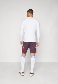 Camiseta atlética blanca de manga larga, pantalones cortos burdeos con acentos rojos, calcetas blancas hasta la rodilla con rayas azules y botas de fútbol negras.