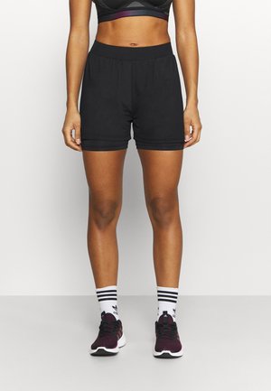 Kvinde iført sorte atletikshorts, sort sports-BH, hvide Adidas-strømper og sorte og lilla løbesko, stående mod en ensfarvet baggrund.