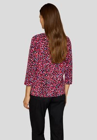 Frau mit langen braunen Haaren, die ein rot-schwarz-lila gemustertes Oberteil mit Dreiviertelärmeln und eine schwarze Hose trägt, mit dem Rücken zur Kamera.