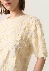Cremefarbene Bluse mit kurzen Ärmeln, die mit komplizierter floraler Stickerei, einer strukturierten Oberfläche und einem Rundhalsausschnitt versehen ist.