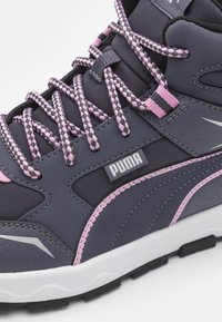 Chaussure de sport synthétique grise avec des accents roses et noirs, surface texturée et semelle intermédiaire blanche contrastante. Présente des lacets tressés et un détail de logo.