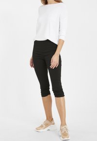 Černé krátké legíny s hladkou texturou, doplněné volným bílým dlouhým rukávem. Oblečení dokončují béžové sportovní tenisky.